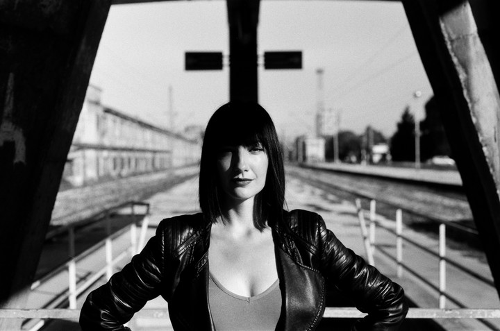A woman with dark hair and a leather jacket stands on a train platform.