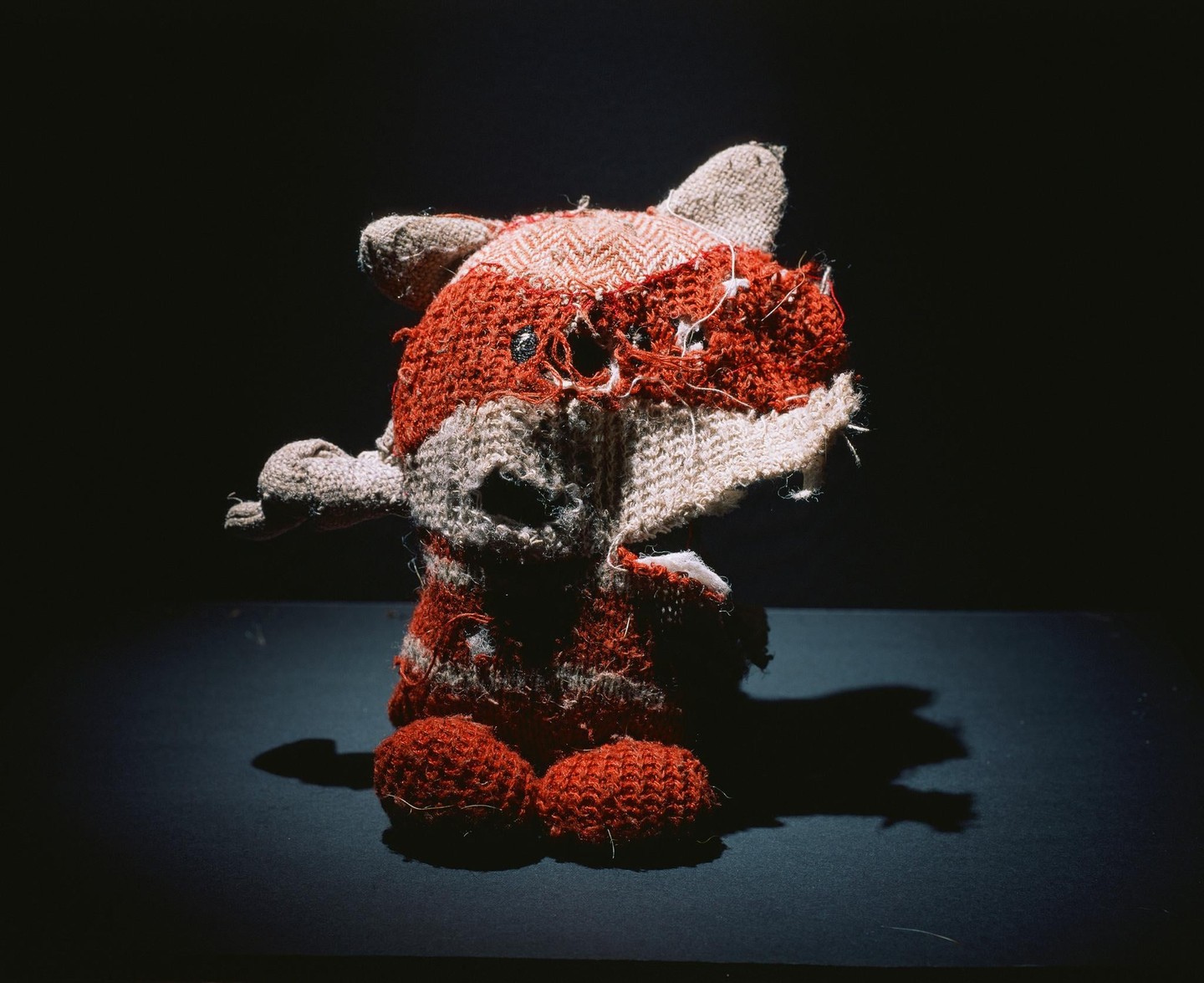A worn, handmade fox toy with red and beige fabric sits on a dark surface, illuminated by a spotlight.