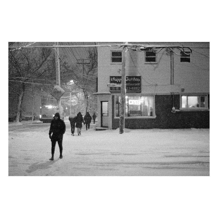 People walk on a snow-covered street past a restaurant called Happy Garden at night.