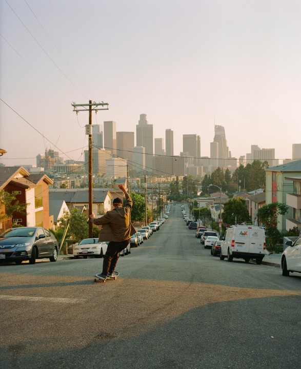 A man on a skateboard rides down a residential street towards a city skyline at dusk.