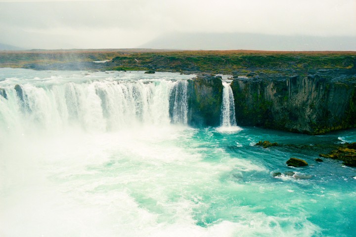 A powerful waterfall cascades into a turquoise river surrounded by rocky cliffs and green landscape.