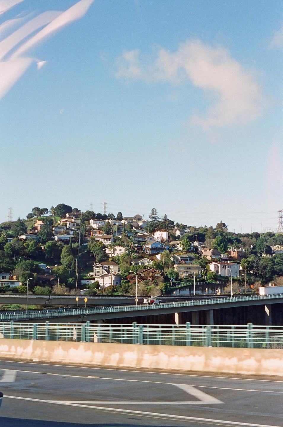 A hillside neighborhood with many houses and trees overlooks a highway and bridge.