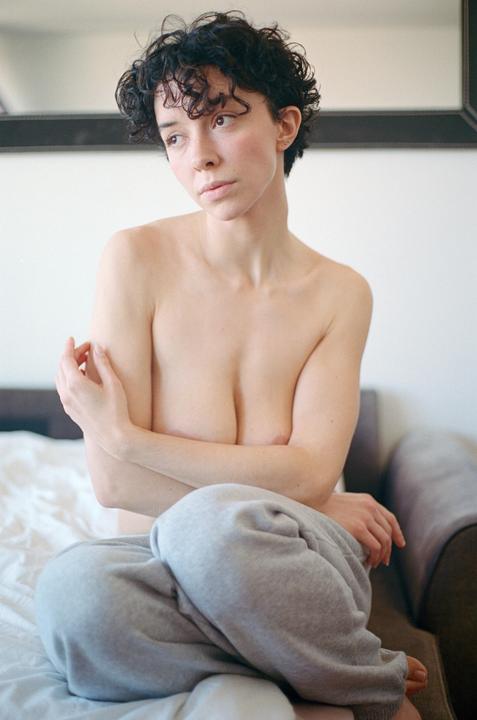 A woman with short curly hair sits topless with her arms crossed, wearing gray sweatpants.
