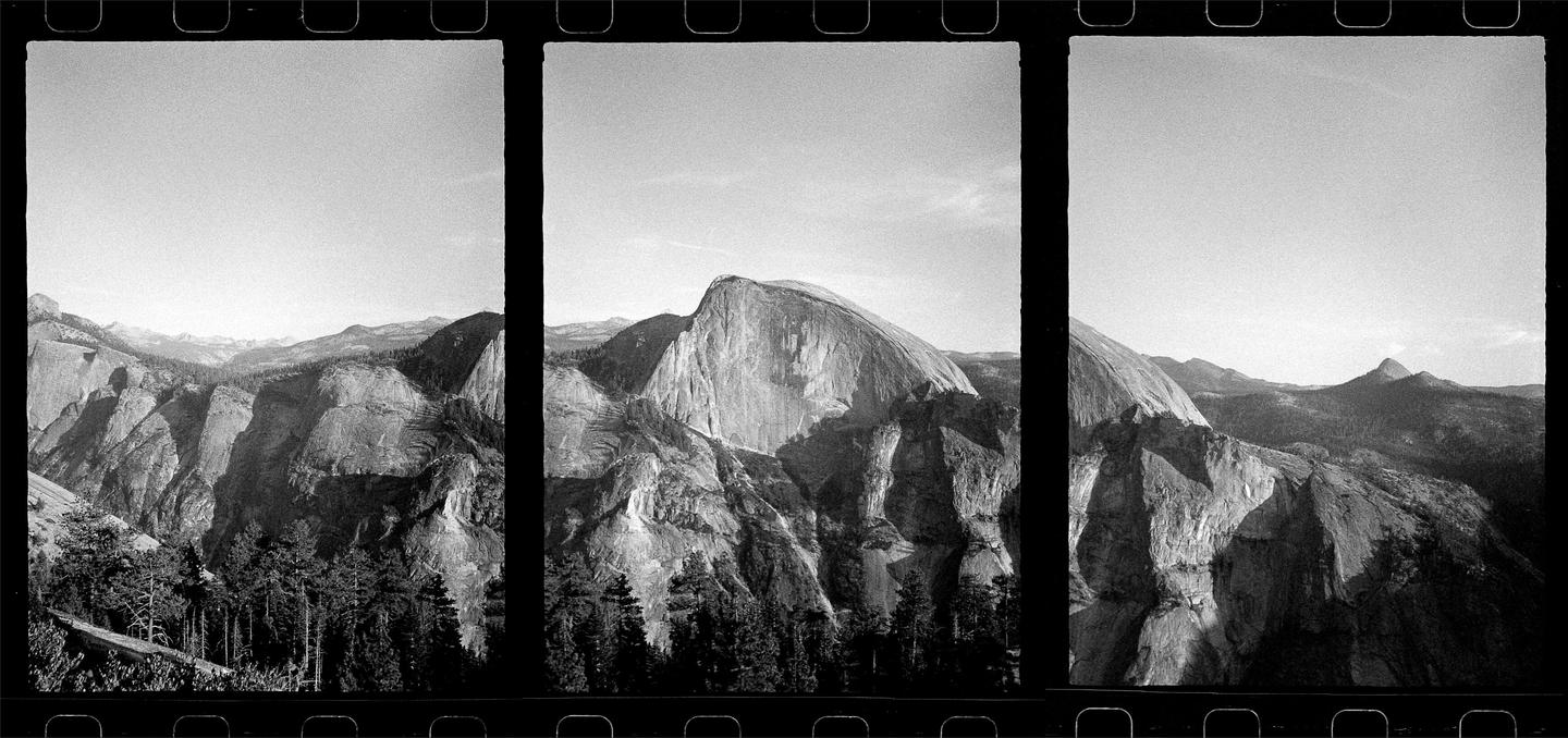 A panoramic black and white landscape shows a large rock formation with a rounded top, surrounded by other mountains and a forest.