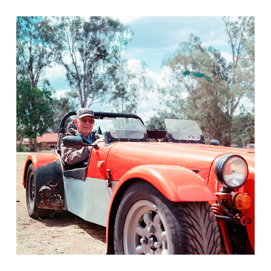 A man in a cap and sunglasses sits in a red and gray convertible car.