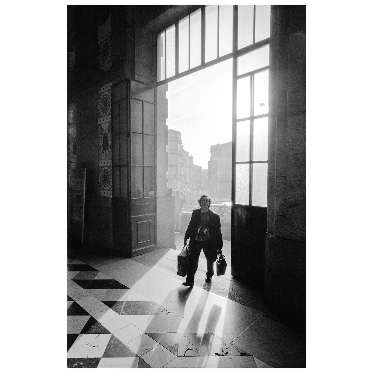 A woman carrying two bags walks out of a building into a bright street.