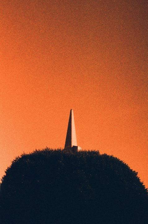 A white obelisk rises from a dark, rounded bush against an orange sky.