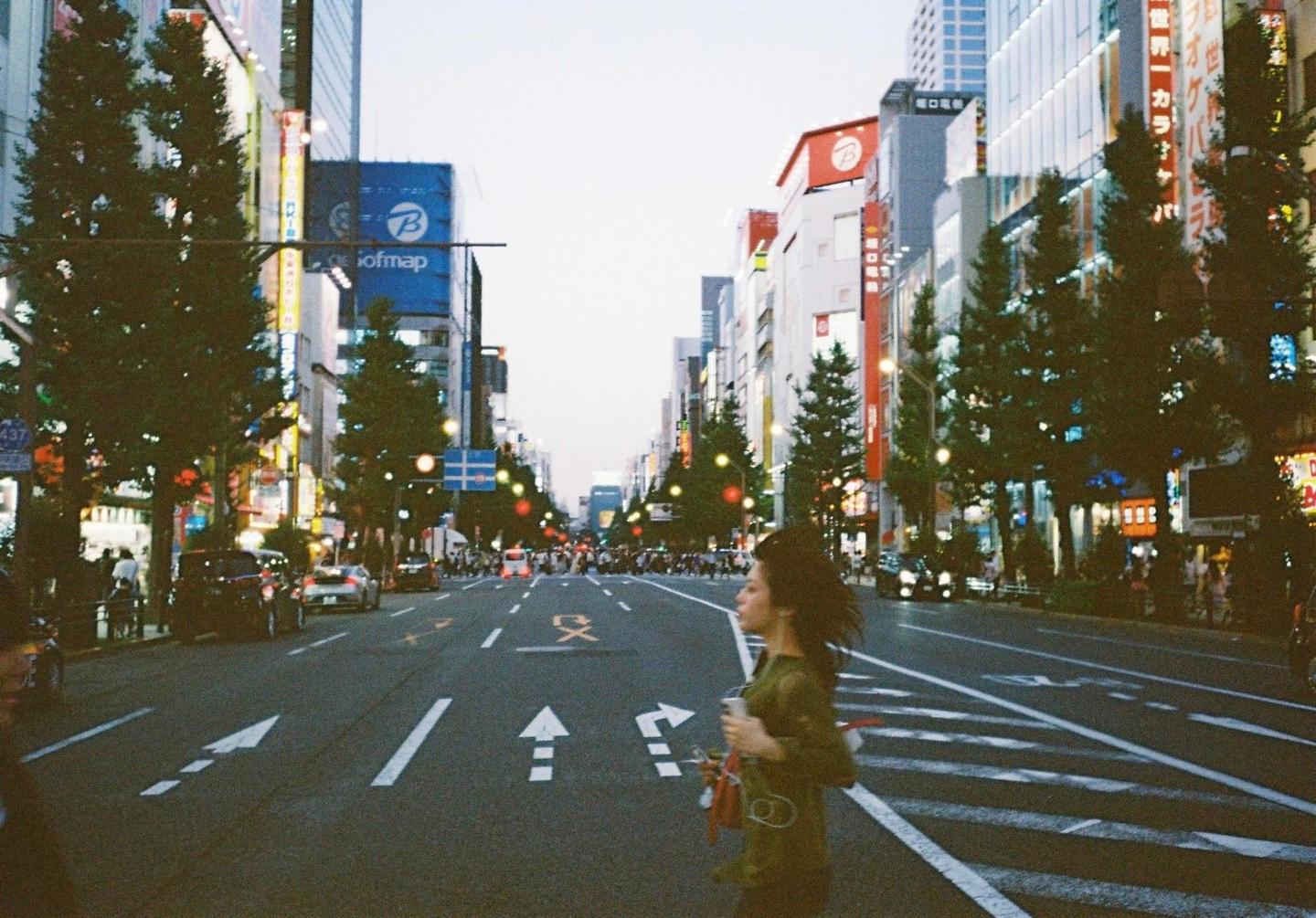 A woman crosses a wide street lined with tall buildings and trees in a city at dusk.