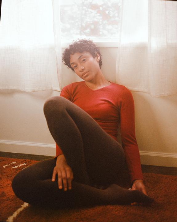 A woman with curly hair sits on a red rug next to a window with sheer white curtains.