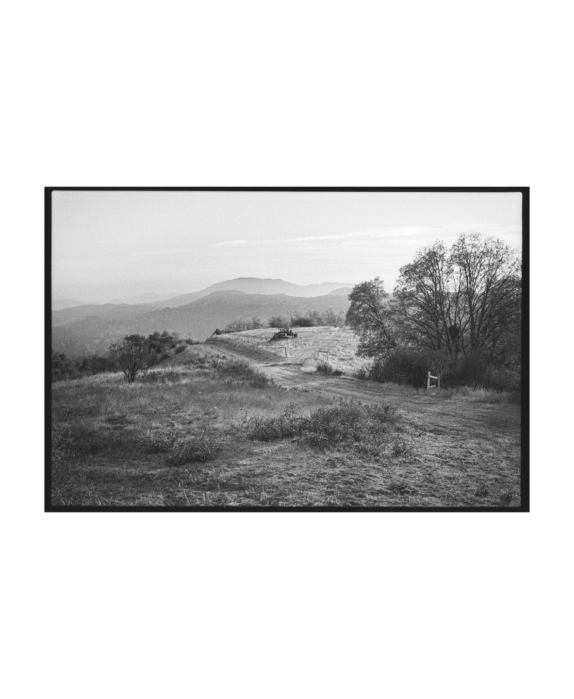A black and white landscape shows rolling hills and mountains under a hazy sky, with a dirt path leading through a grassy field.