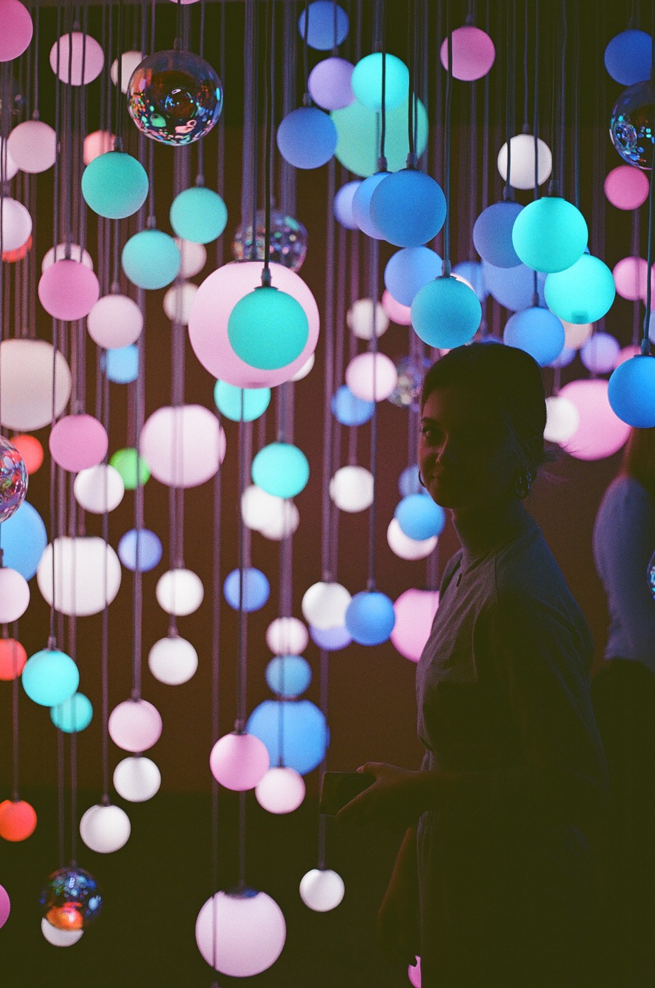 A woman stands in a dark room filled with hanging colorful glowing orbs.