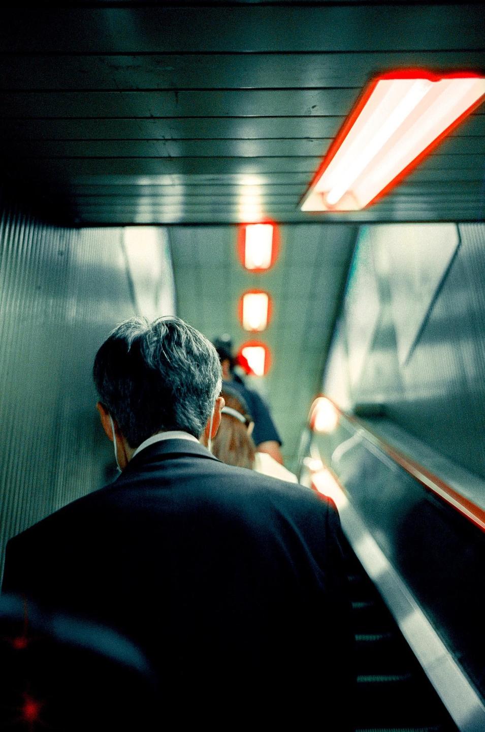 A man in a suit ascends an escalator with red lights on the ceiling.