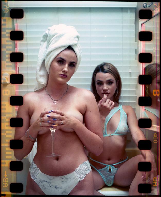 Two women pose in lingerie, one holding a wine glass and the other sitting.
