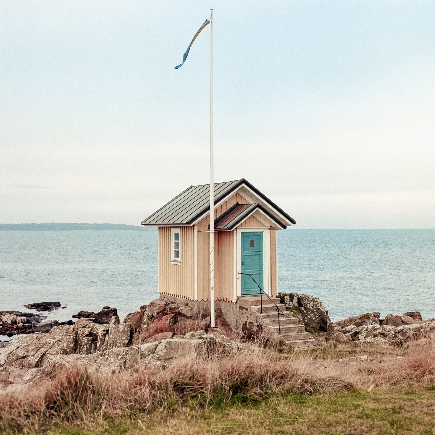 A small wooden house with a blue door stands on a rocky shore next to the sea, with a flag flying from a pole.