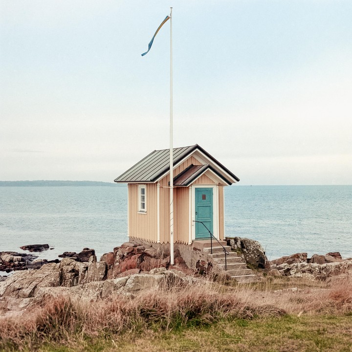 A small wooden house with a blue door stands on a rocky shore next to the sea, with a flag flying from a pole.