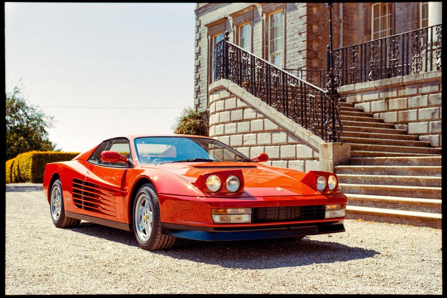 A red Ferrari Testarossa is parked on a gravel driveway in front of a large stone building with an ornate staircase.