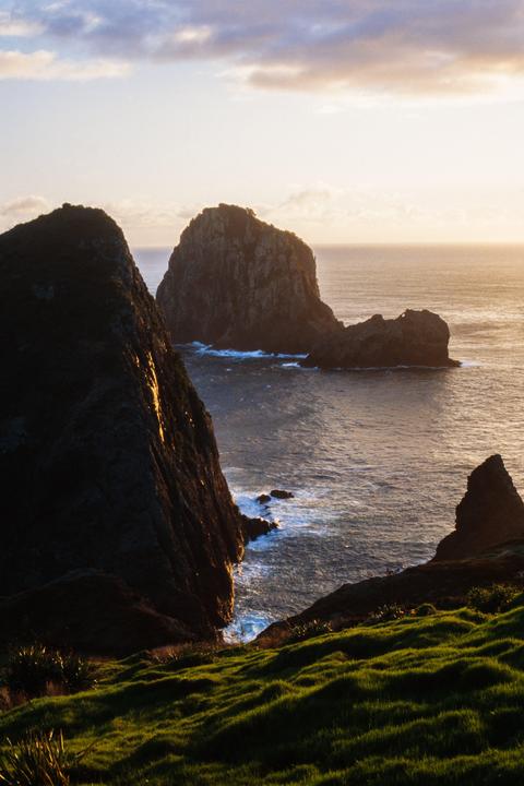 The sun rises over the ocean, illuminating a rocky coastline with a large sea stack in the distance.