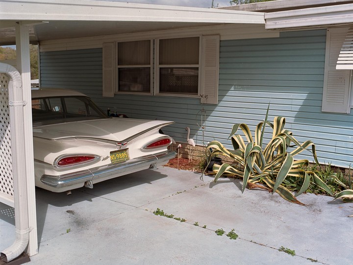 A white car with prominent tail fins is parked under a carport next to a house with blue siding and a large agave plant.