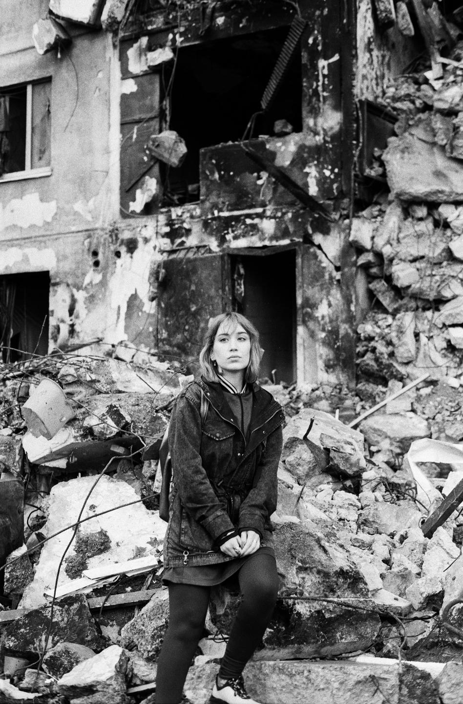A woman stands in front of a destroyed building with rubble all around her.