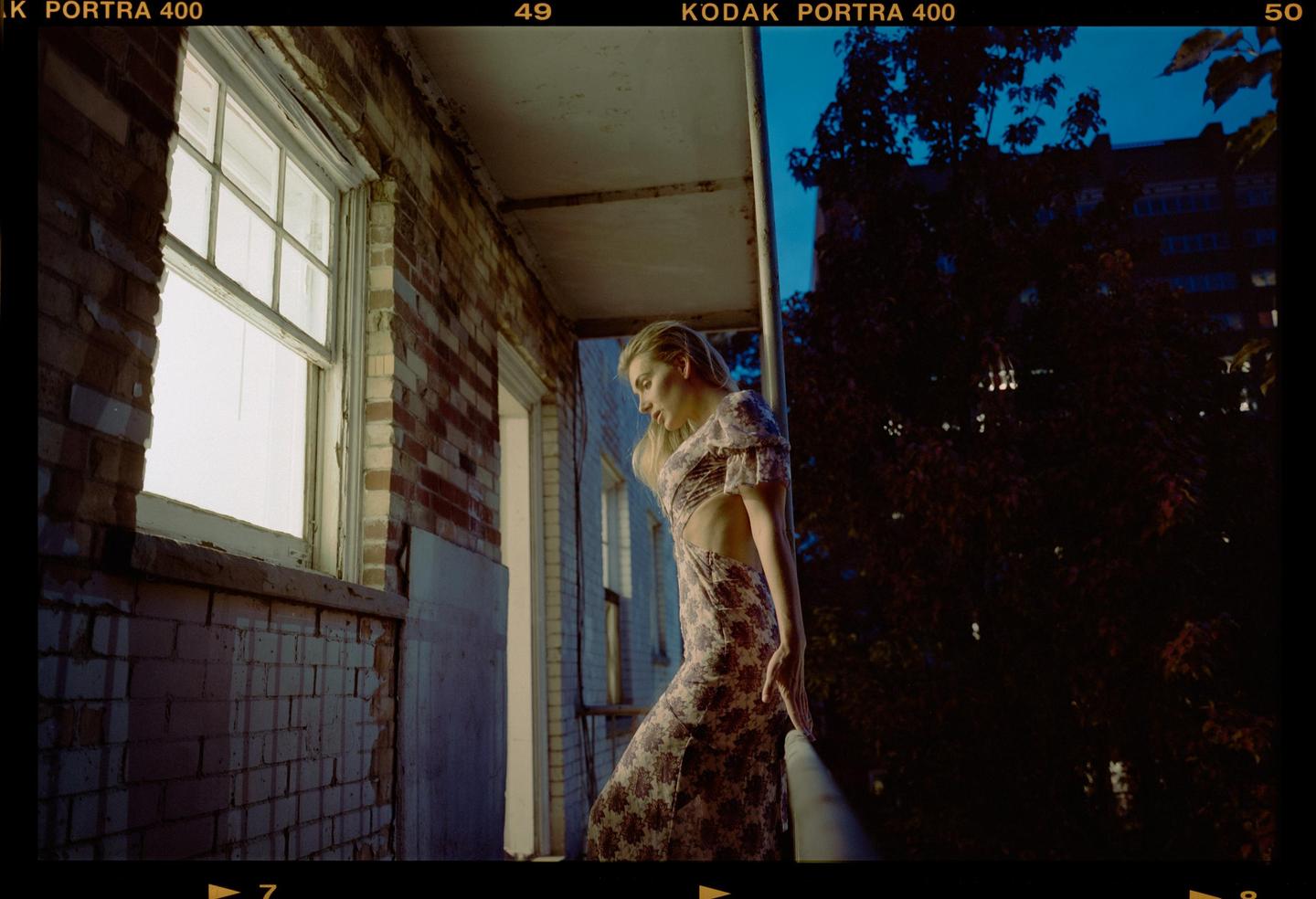 A woman in a floral dress stands on a balcony next to a brick wall and window during blue hour.