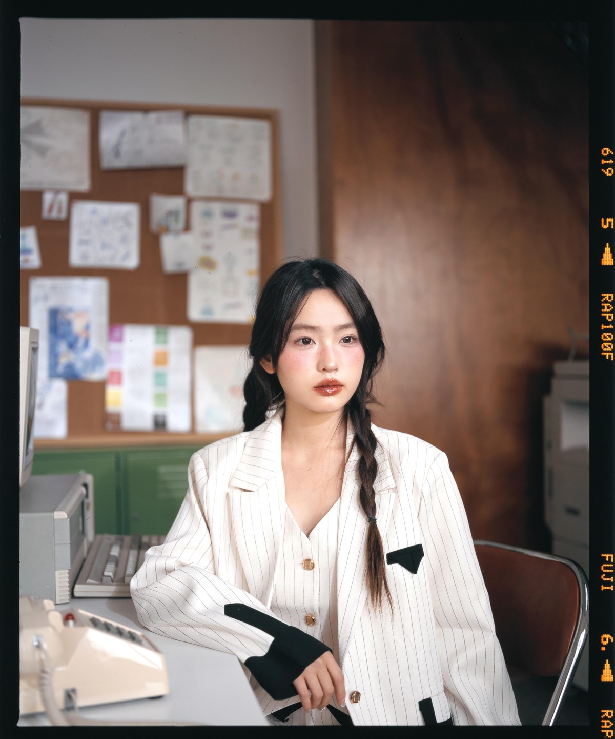 A woman with braided hair sits at a desk in an office, wearing a pinstriped blazer.