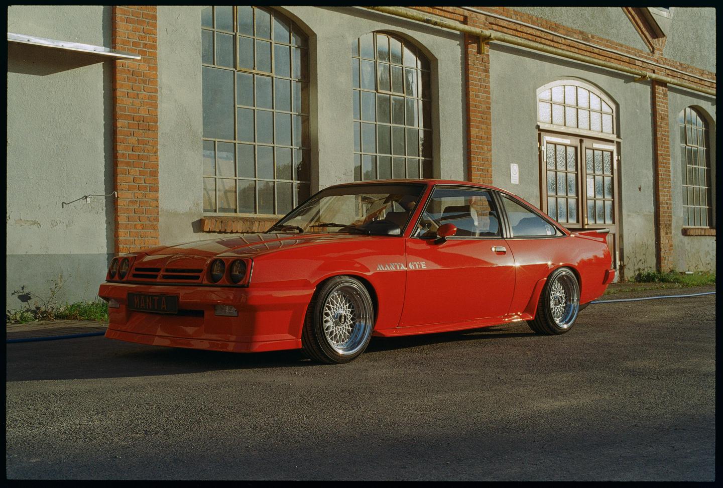 A red Opel Manta GT/E is parked in front of a building with large arched windows.
