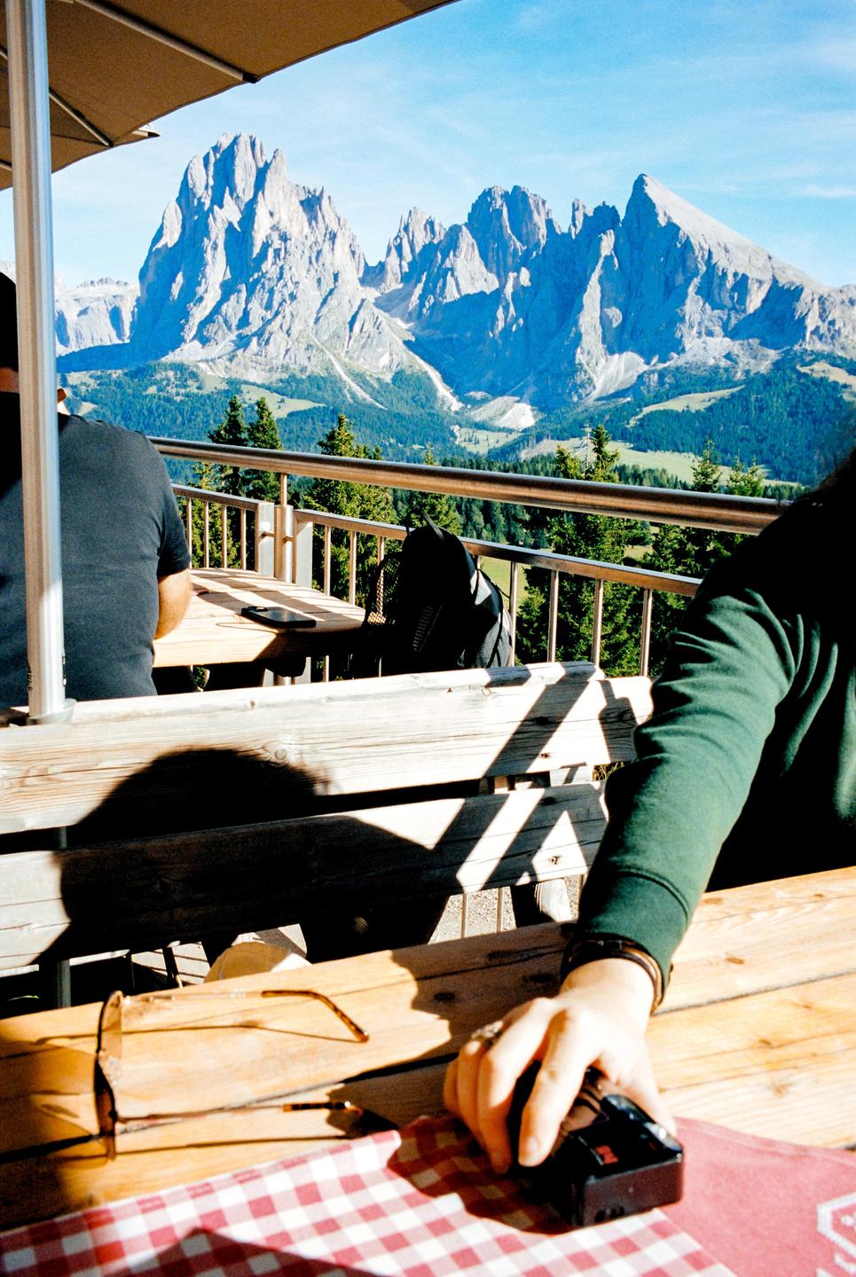 A person sits at a wooden table with a red and white checkered tablecloth, holding a camera, with mountains in the background.