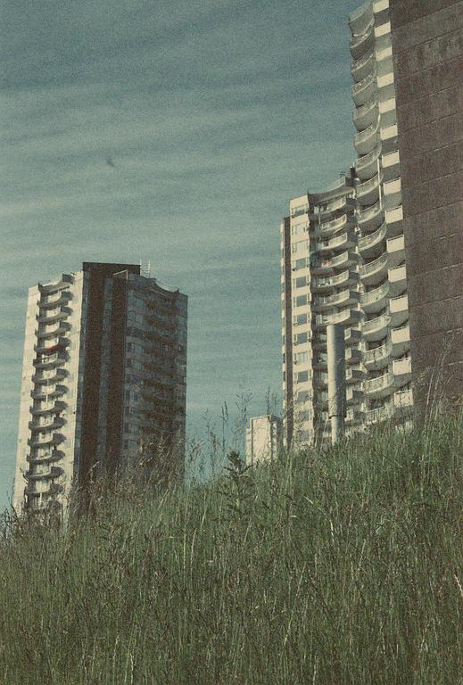 Two tall apartment buildings rise above a field of tall grass under a cloudy sky.