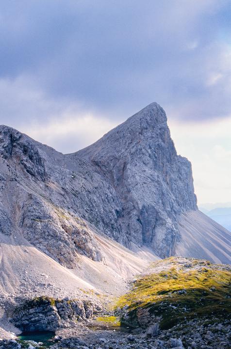 A large, jagged mountain peak rises under a cloudy sky, with a small body of water and green moss in the foreground.