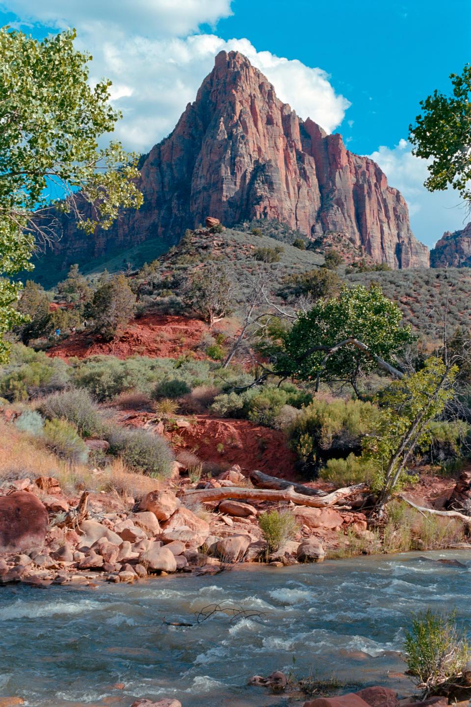 A river flows past a rocky bank with trees and shrubs, leading up to a large red mountain under a blue sky with clouds.