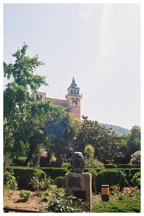 A stone bust on a pedestal stands in a lush garden with a church tower visible in the background.