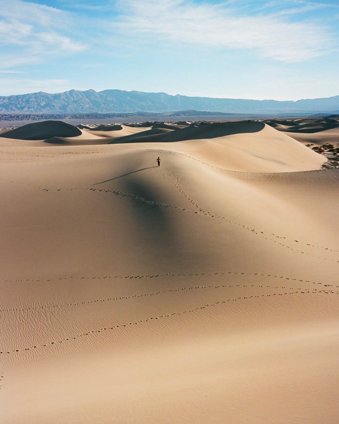 A person walks across a sand dune with mountains in the background.
