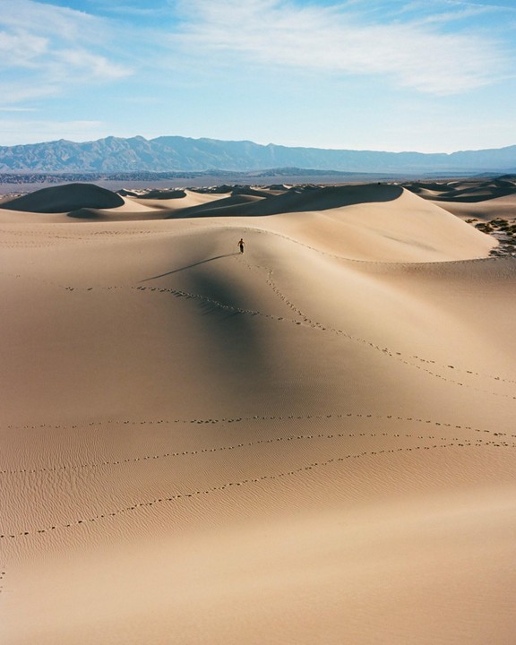 A person walks across a sand dune with mountains in the background.