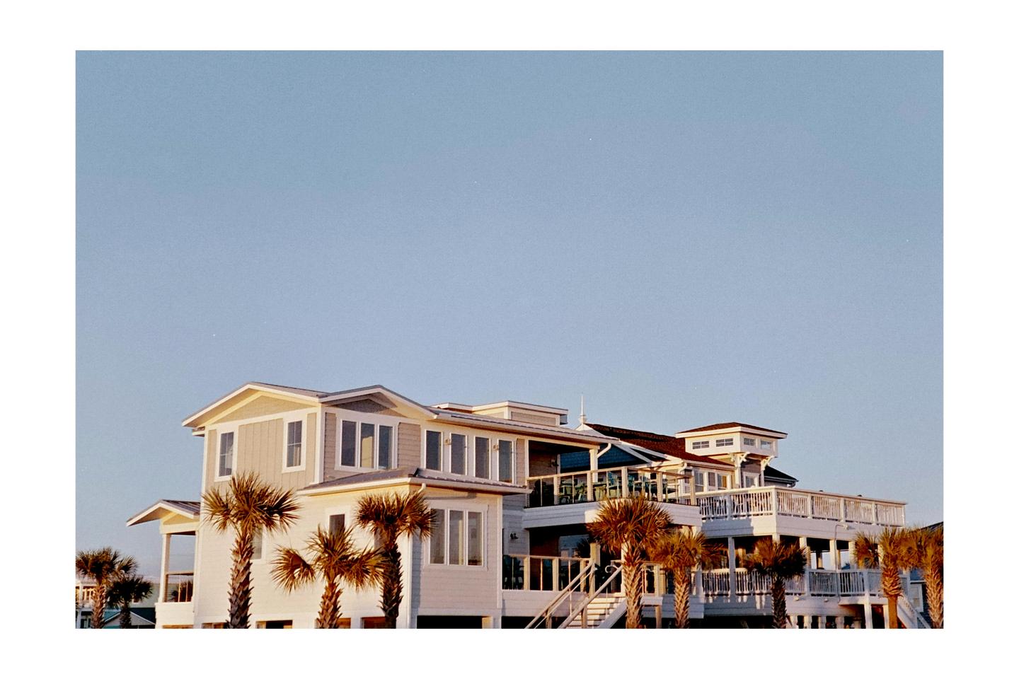 A large beach house with multiple balconies and palm trees stands under a clear sky.