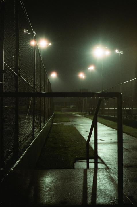 A wet path next to a chain link fence is illuminated by several bright lights at night.