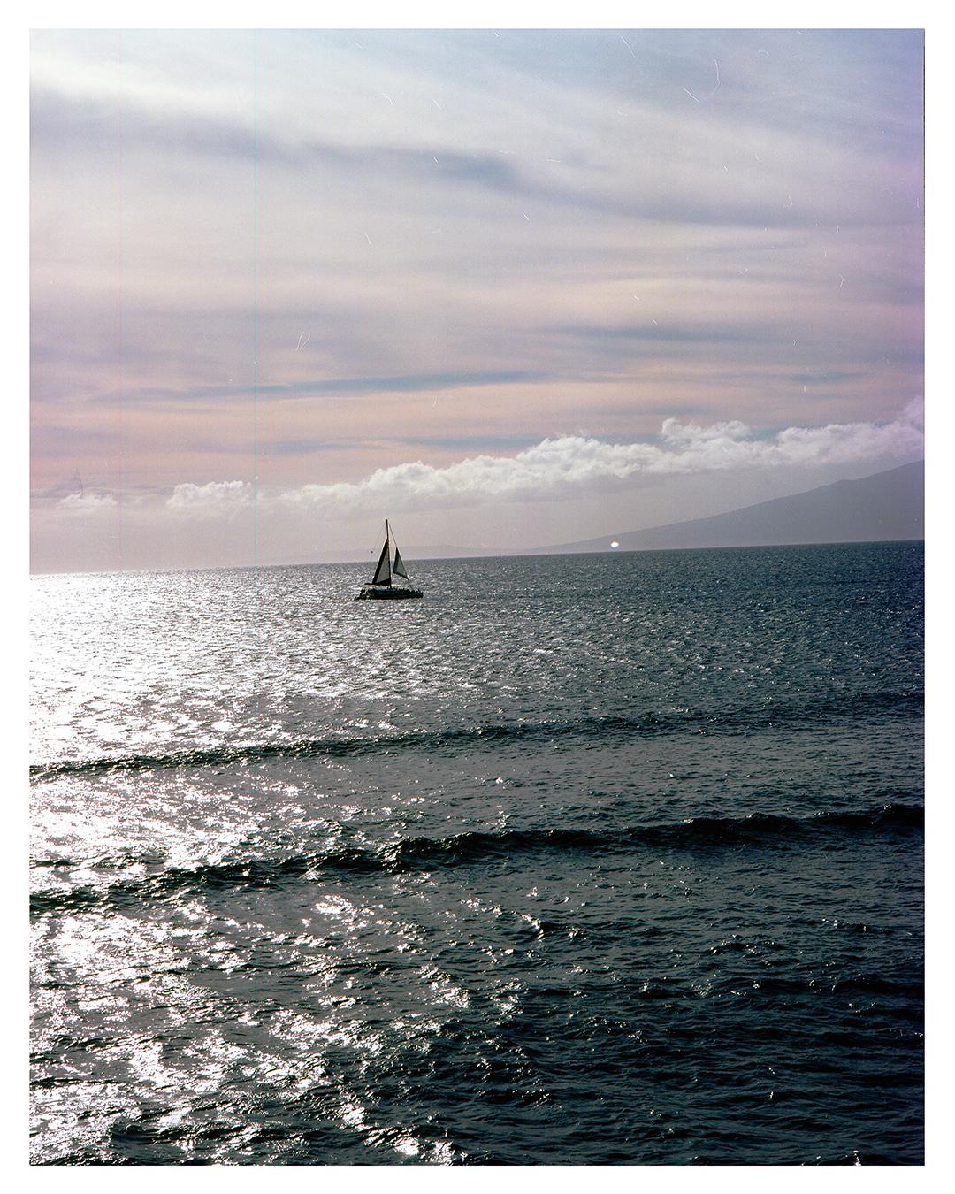 A sailboat is visible on the ocean with a mountain in the distance under a cloudy sky.