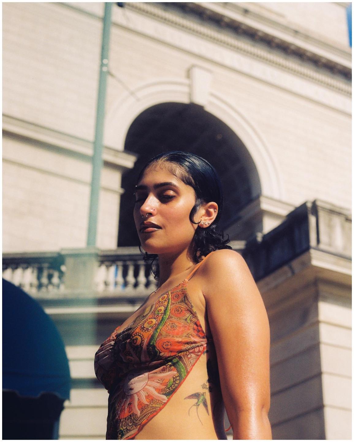 A woman with closed eyes and a patterned top stands in front of a building with an arched window.