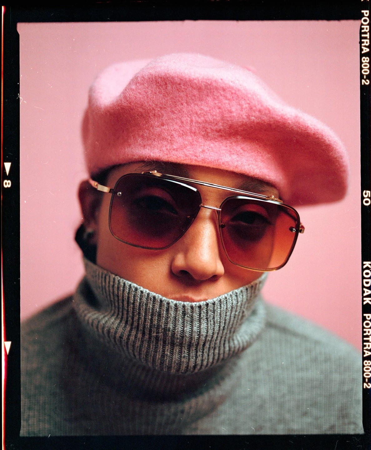 A person wearing a pink beret, sunglasses, and a grey turtleneck sweater poses against a pink background.
