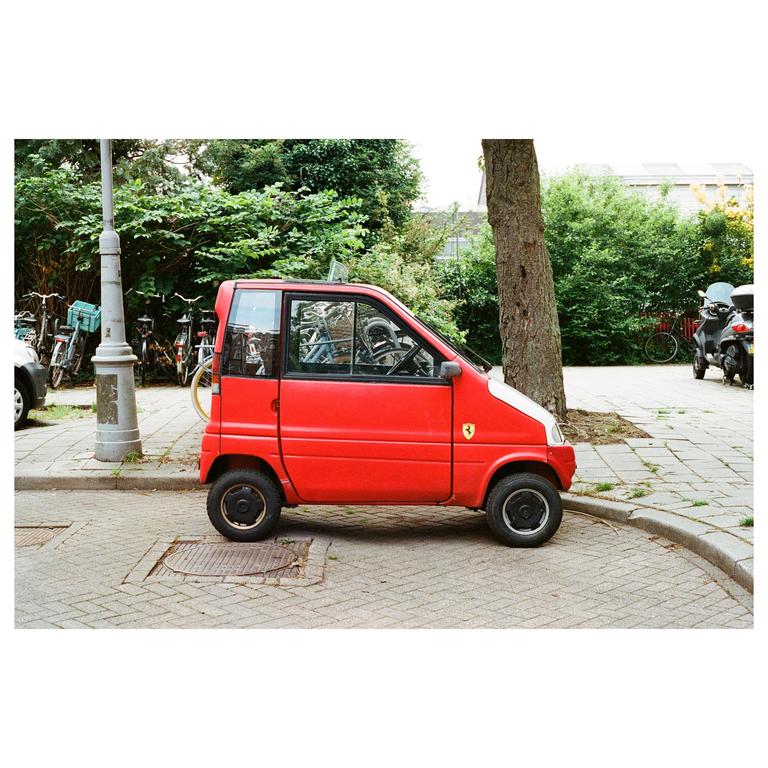 A small red car is parked on a paved street next to a tree and a lamppost.