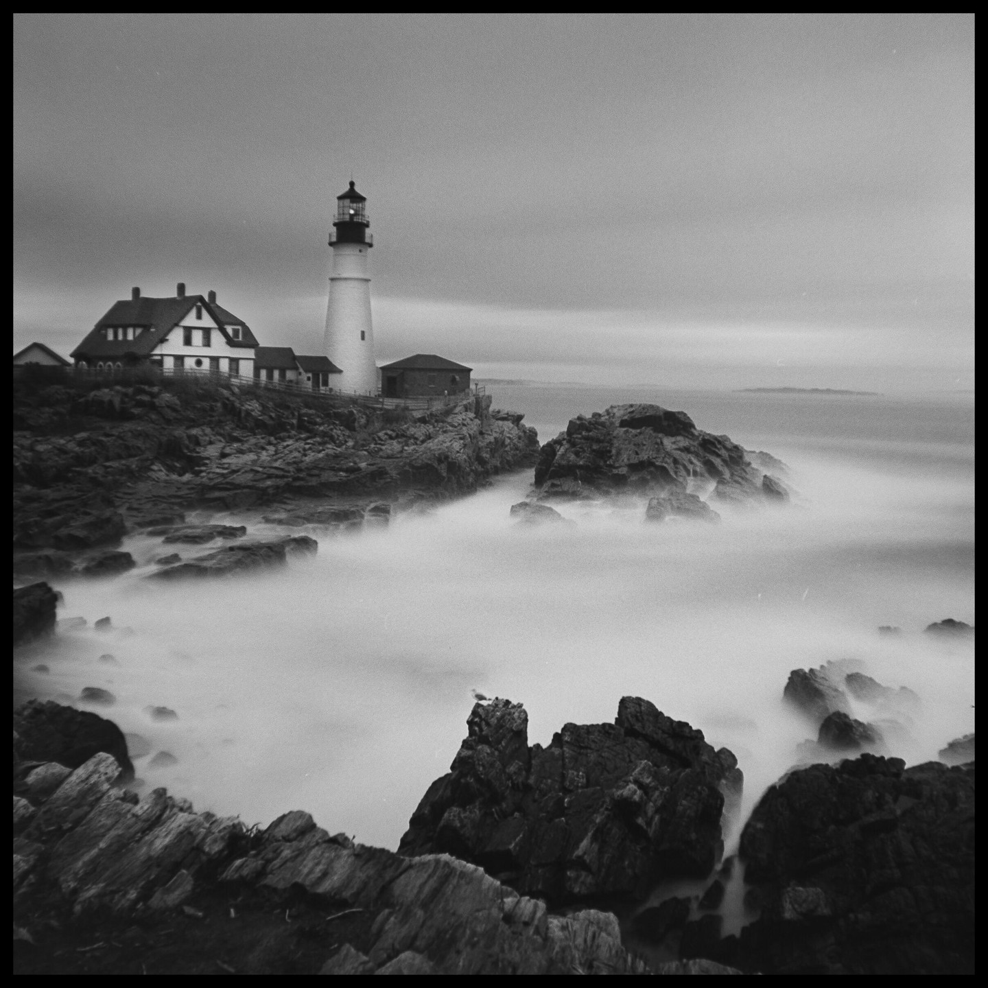 A lighthouse stands on a rocky coast with a house nearby, and the ocean water is smoothed by a long exposure.