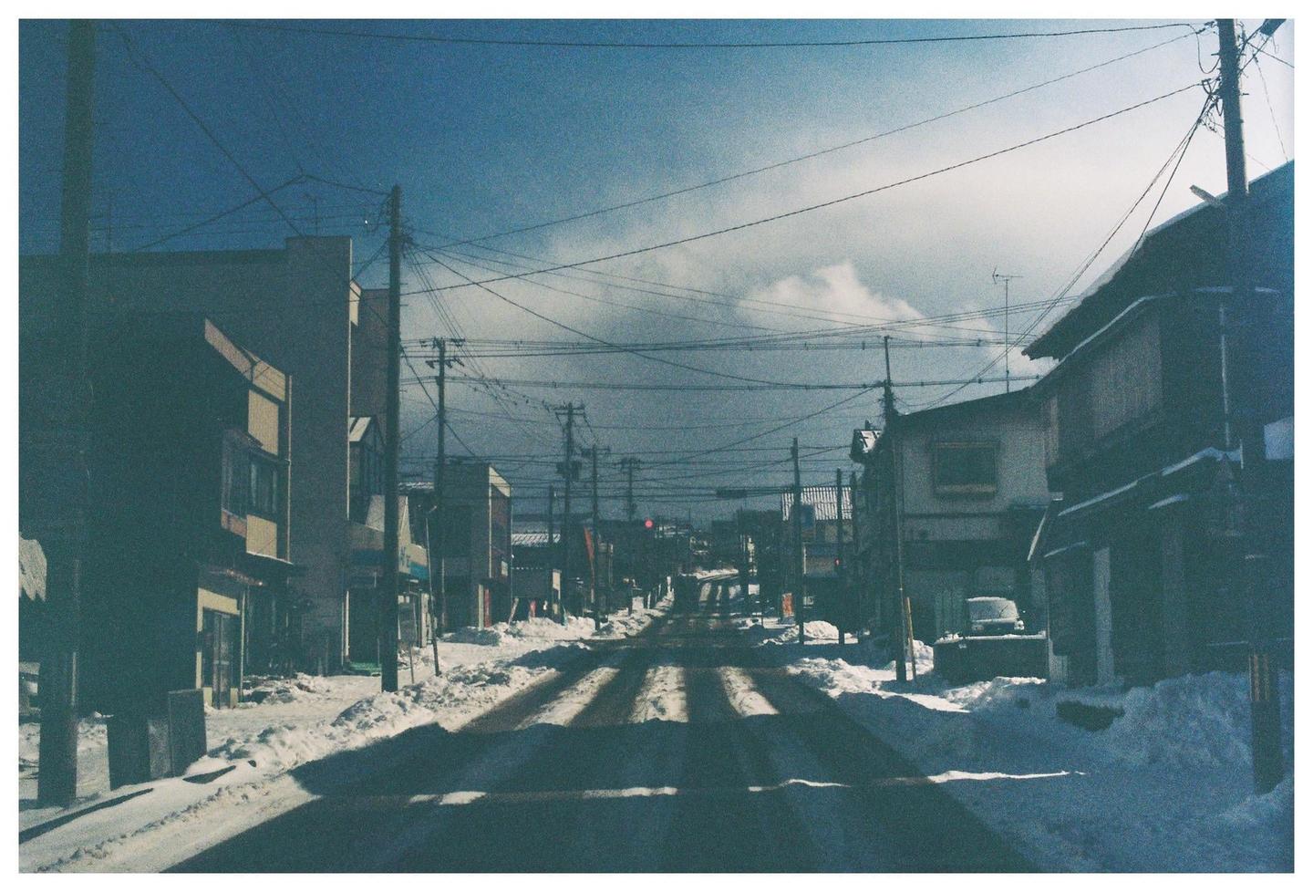 A snow-covered street with buildings and utility poles on both sides stretches into the distance under a cloudy sky.