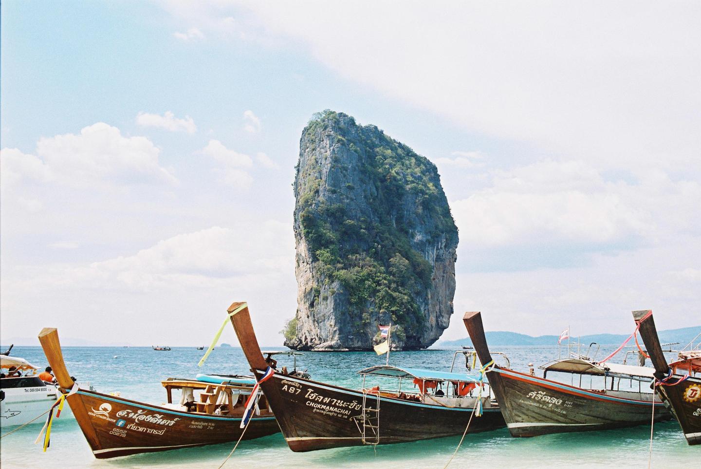 Several longtail boats are moored in shallow turquoise water with a large, tree-covered rock formation in the background under a partly cloudy sky.
