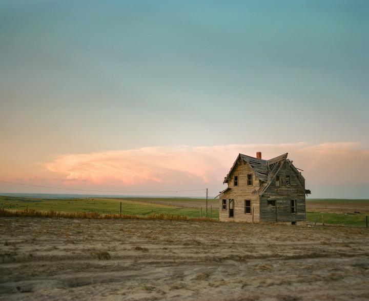 An abandoned wooden house with a damaged roof stands in a field under a sky with pink clouds.