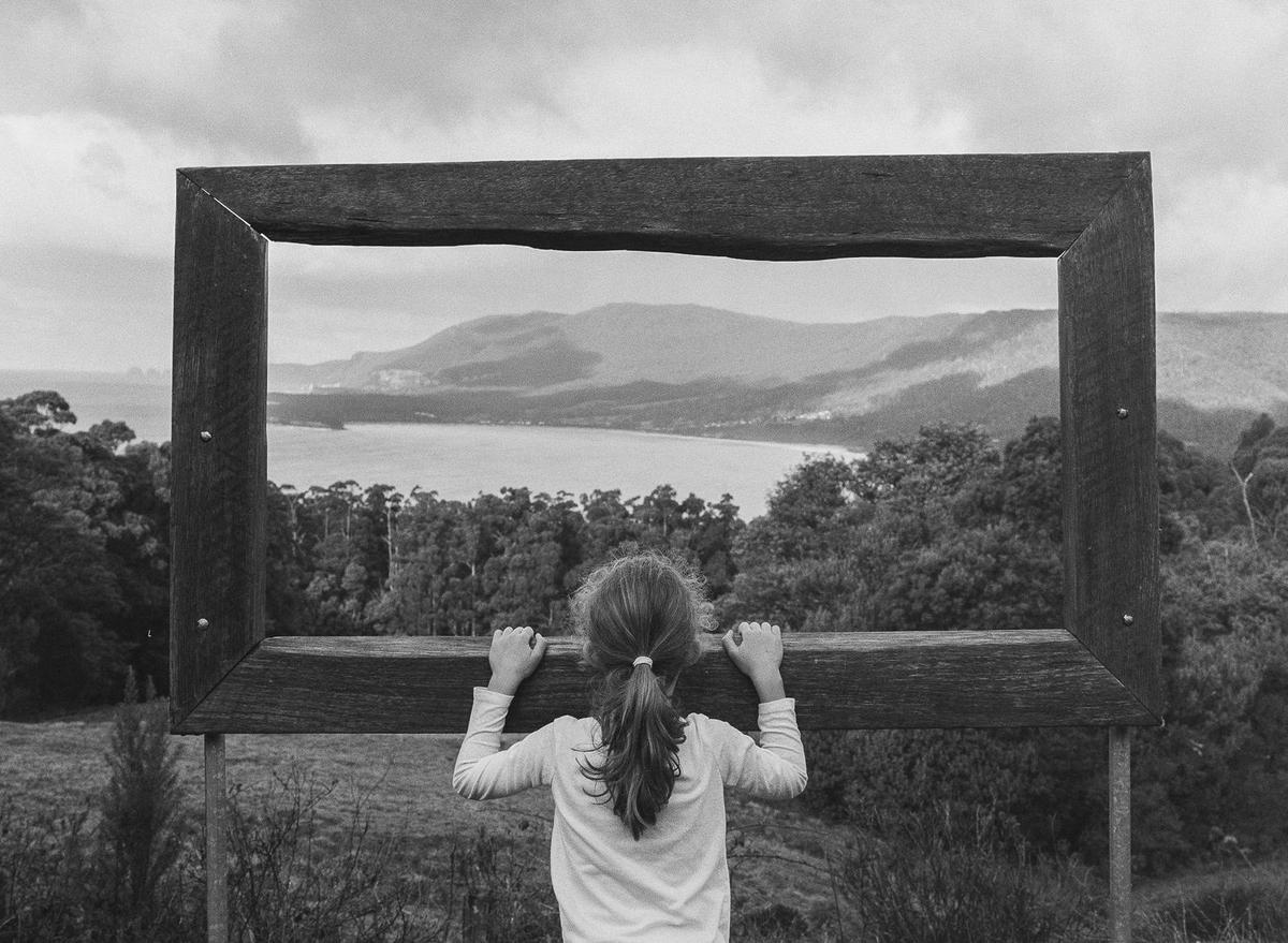 A girl looks through a large wooden frame at a bay surrounded by mountains and trees.