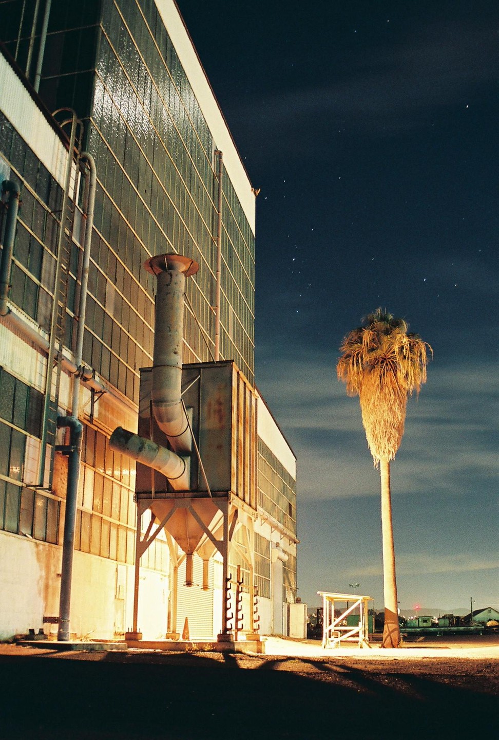 A tall palm tree stands next to a large industrial building with visible pipes and vents under a starry night sky.