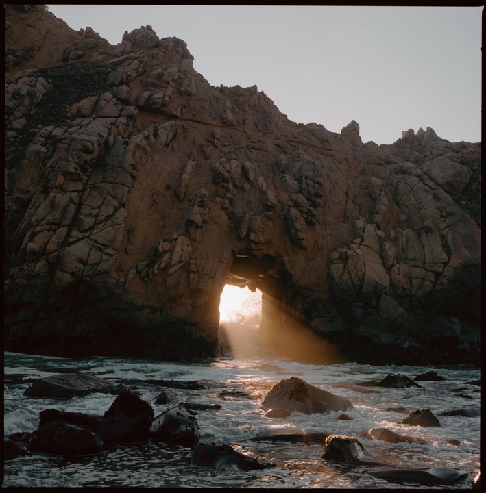 Sunlight streams through a natural arch in a rocky cliff over the ocean.