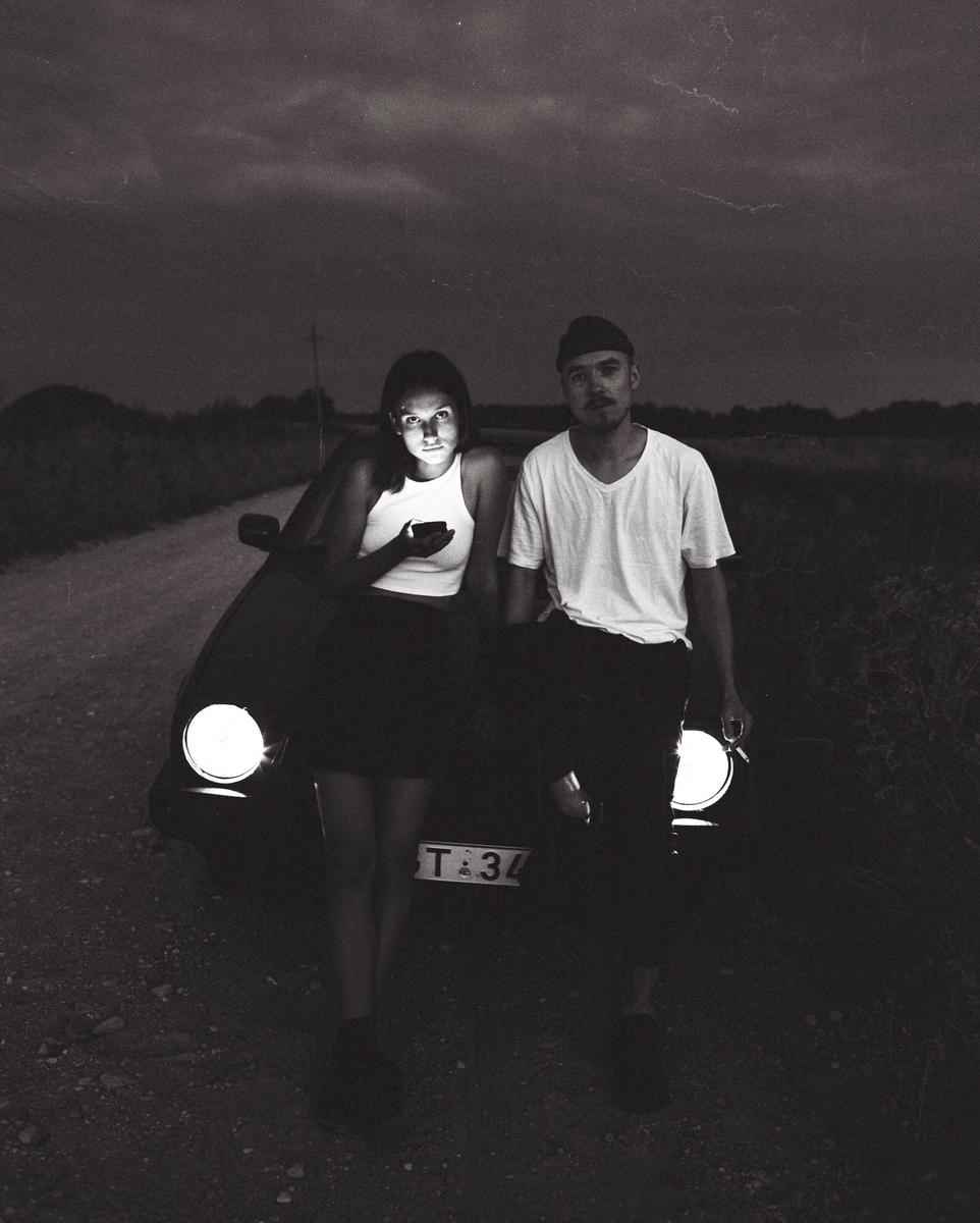 A couple stands in front of a car with its headlights on, with the woman looking at her phone.