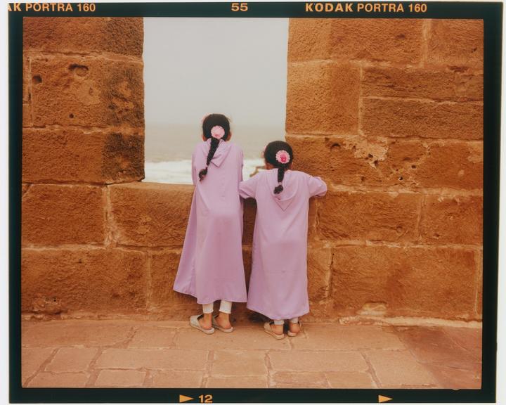 Two girls in purple robes look out through a stone archway at the ocean.