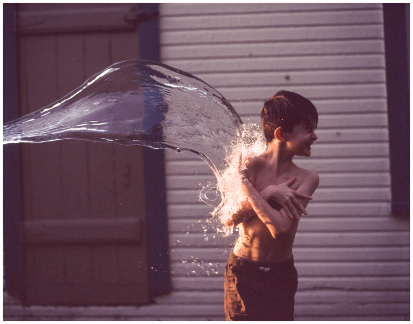 A boy is being splashed with water from the left side of the frame, with a closed door and a white wall in the background.