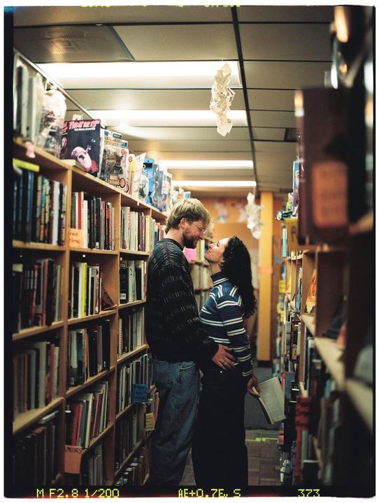A couple stands close together and gazes at each other in a narrow bookstore aisle.